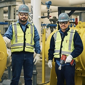 Two men in safety vests standing next to a yellow pipe.