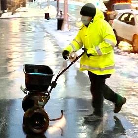 A man in a yellow jacket pushing a snow shovel on a city street.