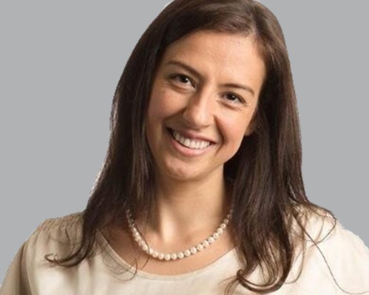 A woman with long brown hair, wearing a white blouse and a pearl necklace, smiling against a gray background.