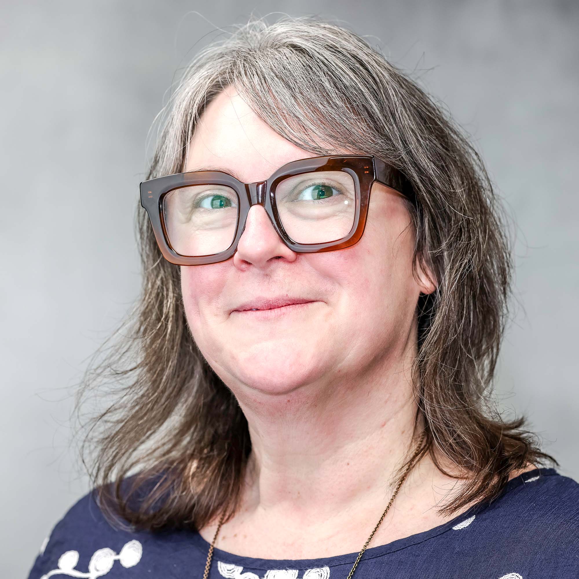 A woman with shoulder-length gray hair, wearing large square glasses and a navy shirt with white patterns, smiles slightly in front of a plain gray background.