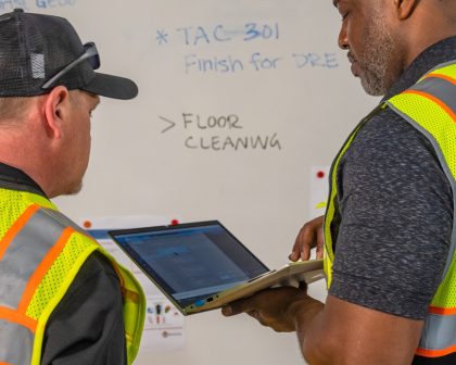 Two men in safety vests looking at a whiteboard.