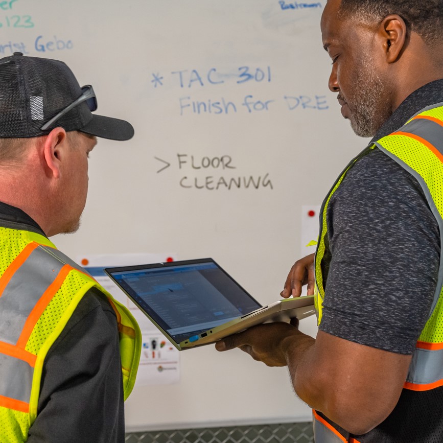 Two men in safety vests looking at a whiteboard.