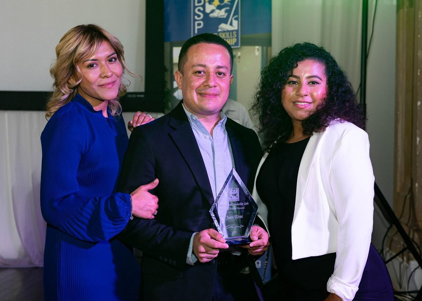 Jaun Rivas holding a crystal award with his name on it, flanked by two women.