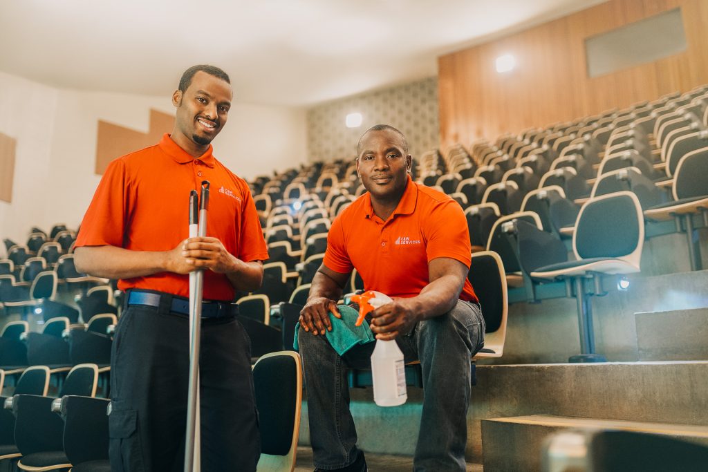 Two men in orange shirts holding a mop and a spray bottle in a auditorium.