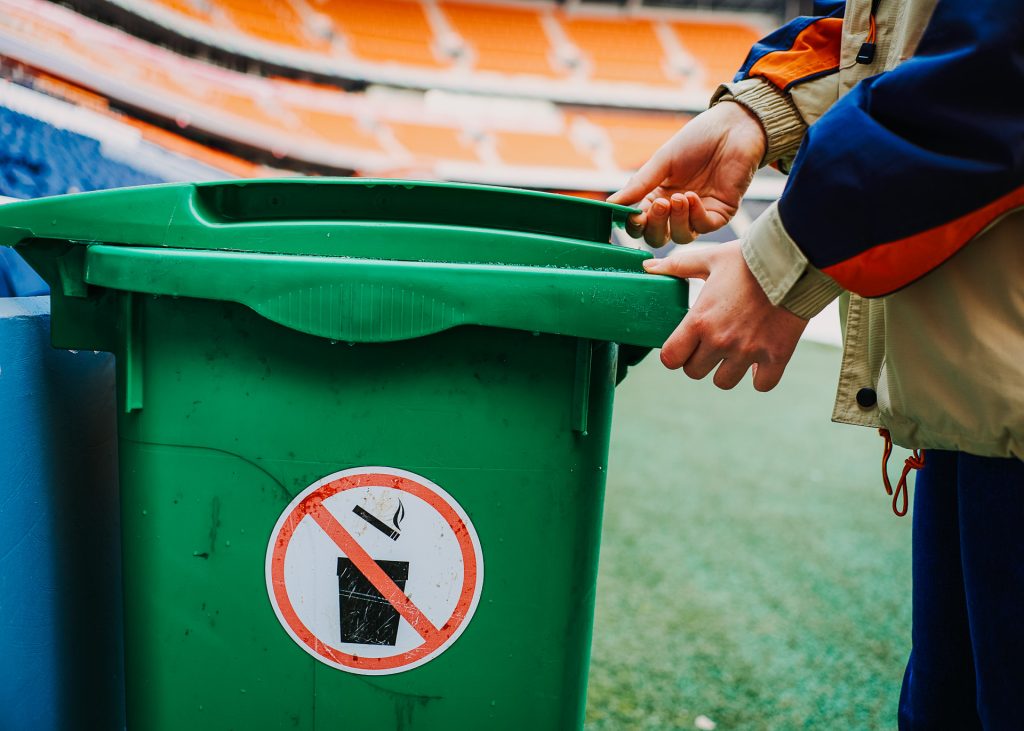 A person putting trash into a green bin with stadium seats in the background.