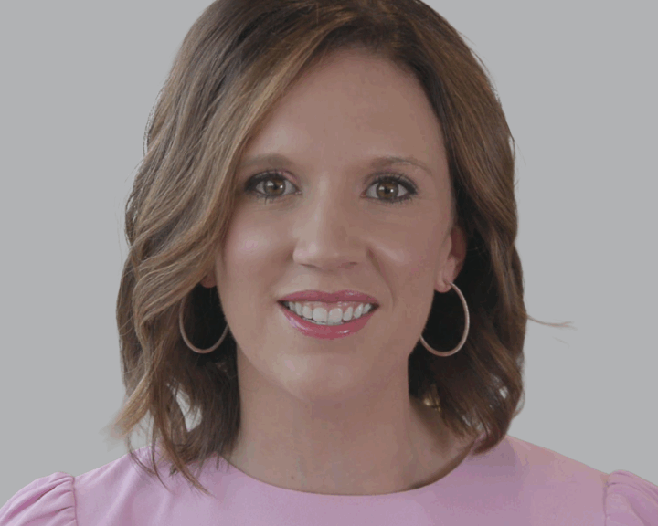 Woman with shoulder-length brown hair wearing hoop earrings and a pink top, smiling in front of a plain light gray background.