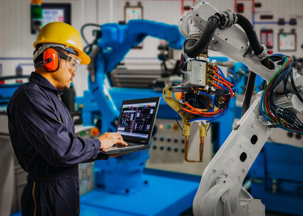 Engineer using laptop for computer maintenance on an automatic robotic hand machine automotive plant.
