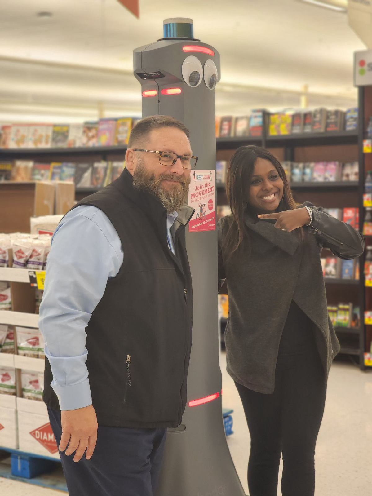 A man and a woman smiling and posing next to a tall, robot-like security device with eyes, inside a supermarket aisle.