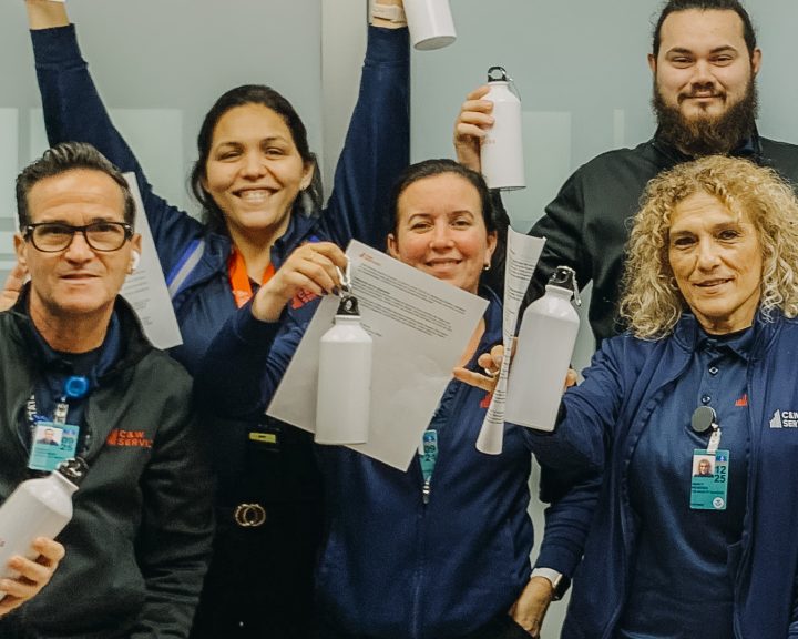Five employees in blue uniforms and lanyards, smiling and holding up white bottles, celebrate in an office setting.