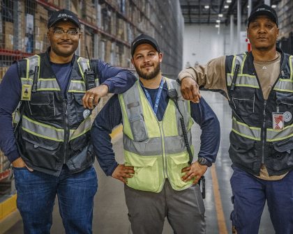 Three warehouse workers in safety vests pose together in a large industrial storage area.