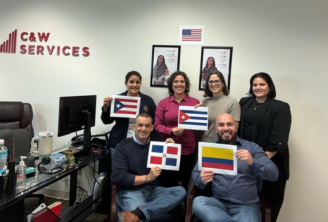 Six people in an office holding flags of various countries. they are smiling and standing in front of a company logo and american flags.