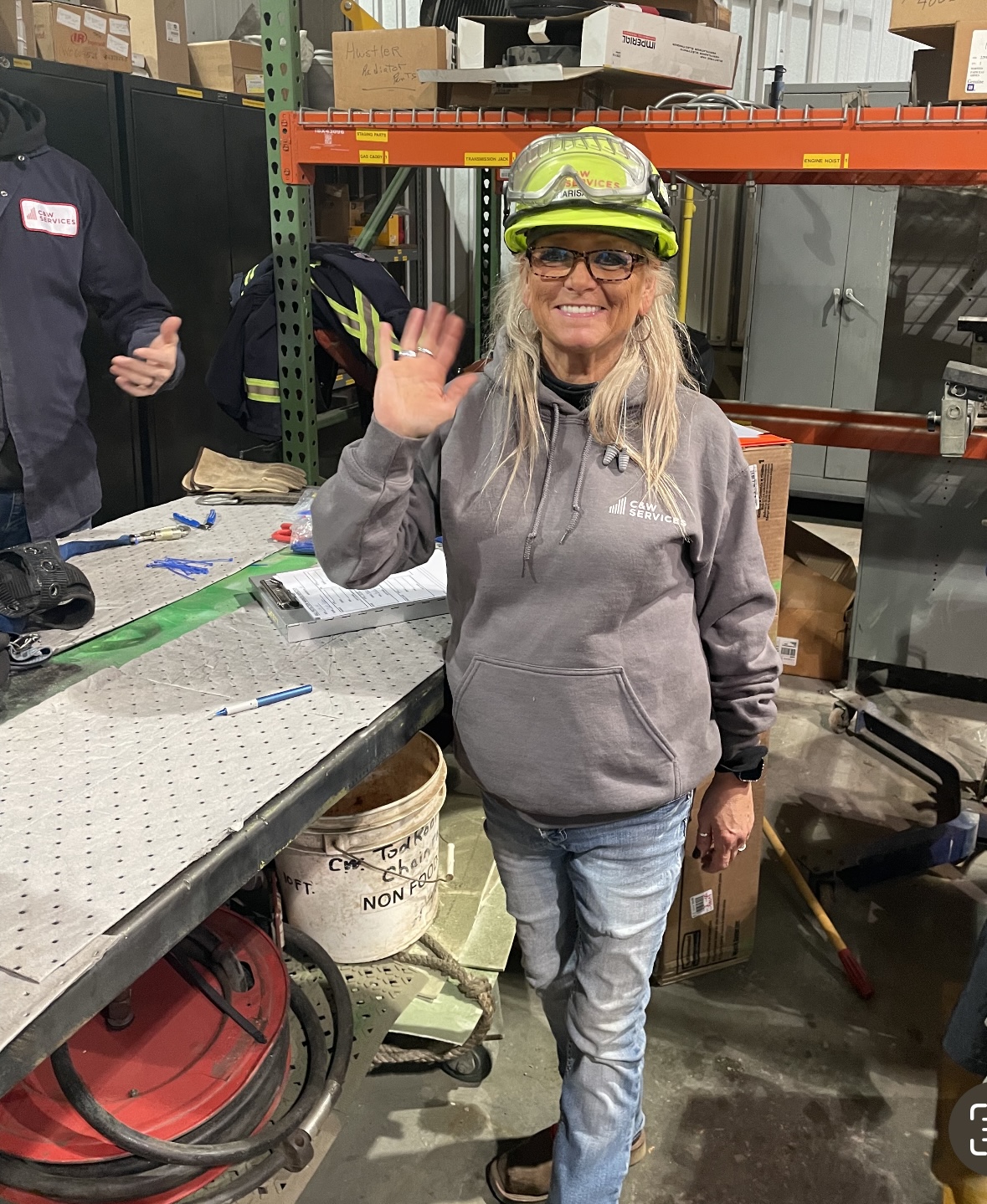 A woman in a workshop wearing a hoodie, safety goggles, and a hard hat with a headlamp is waving at the camera.
