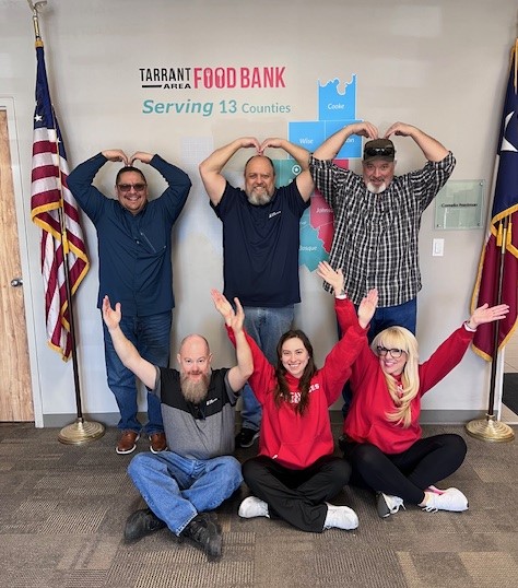 Group of six adults posing with arms raised to form heart shapes above their heads in front of a tarrant food bank sign.
