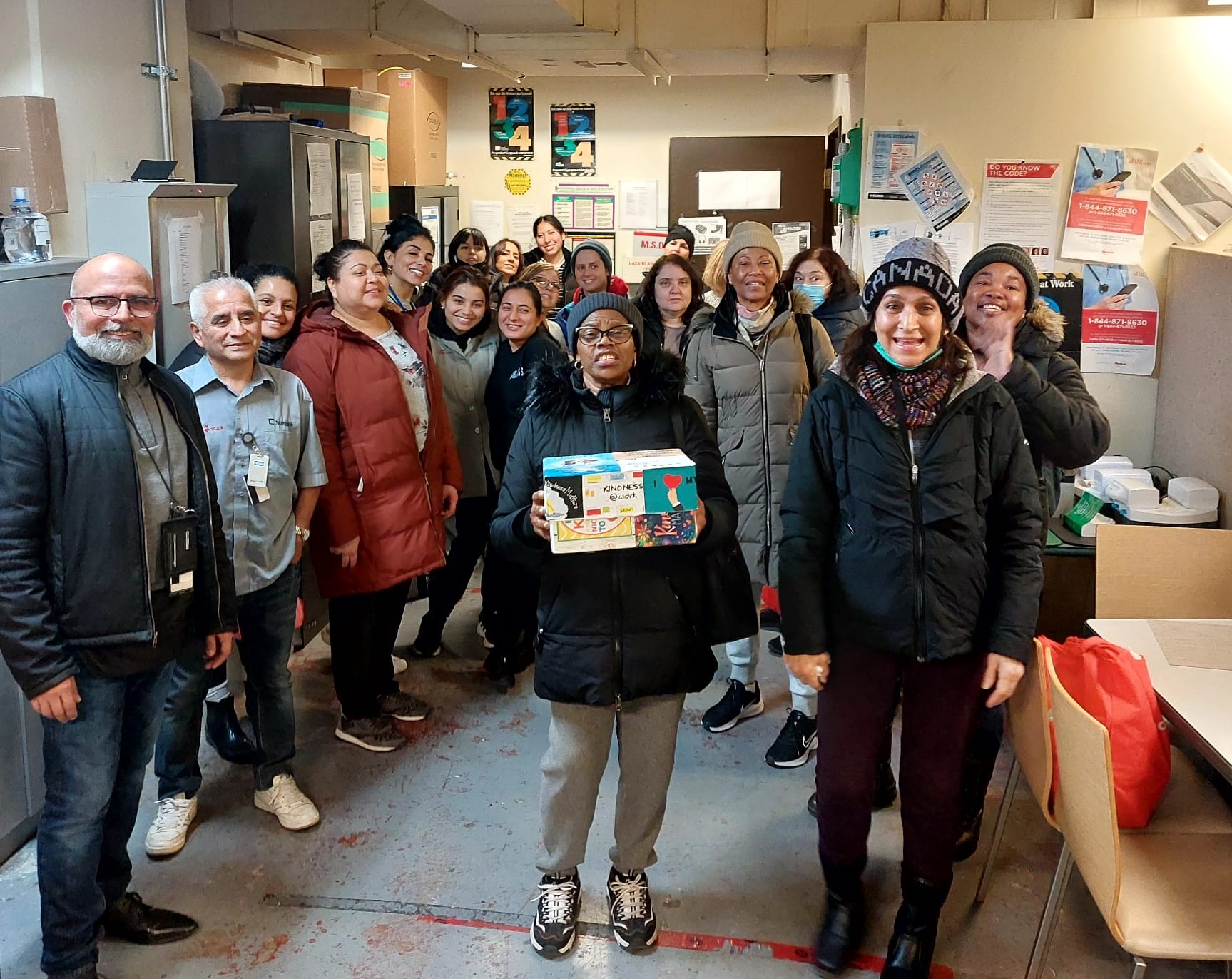 A diverse group of people smiling in a break room, one person holding a box of crayons, posing for a group photo.