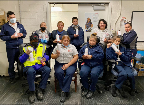 A group of nine diverse custodial staff in uniforms sitting and standing in a break room, each holding cleaning supplies, smiling at the camera.