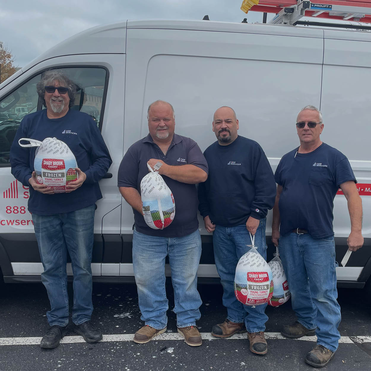 Four men in blue shirts and jeans hold turkeys in front of a white service van in a parking lot.