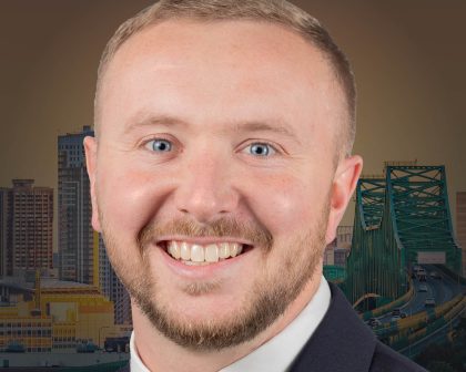 Man smiling in a suit and tie, with a city skyline and green bridge in the background.