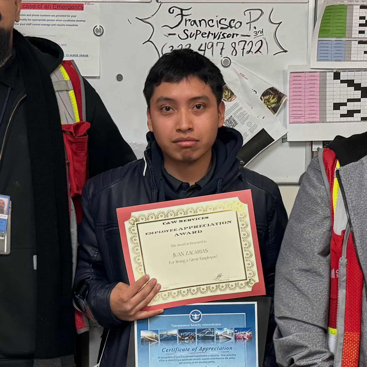 Person holding a "Certificate of Appreciation" and another award, standing in front of a bulletin board with various postings.