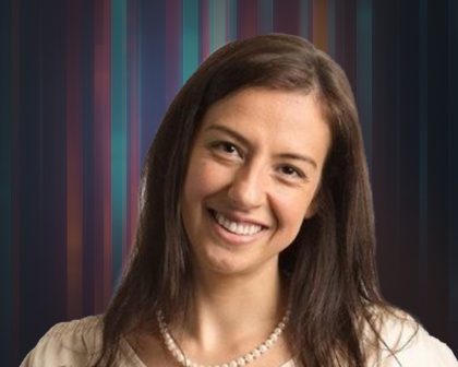 Smiling woman with long brown hair wearing a pearl necklace and beige top, against a striped background.