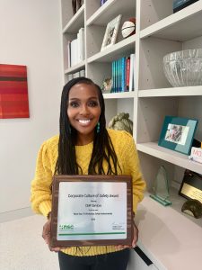 A woman in a yellow sweater smiles while holding a framed Corporate Culture of Safety Award in an office setting with shelves in the background.