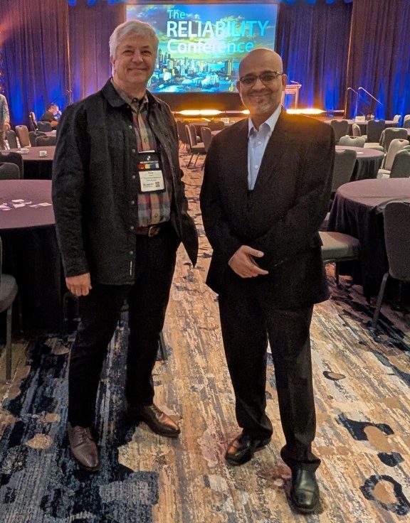 Two men stand and smile for a photo in a conference room with round tables and blue lighting; a stage behind them displays "The Reliability Conference.