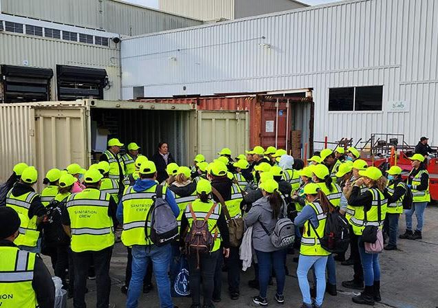 A large group of people in yellow safety vests and caps gathers outside an industrial building, listening to a person speaking near a shipping container.