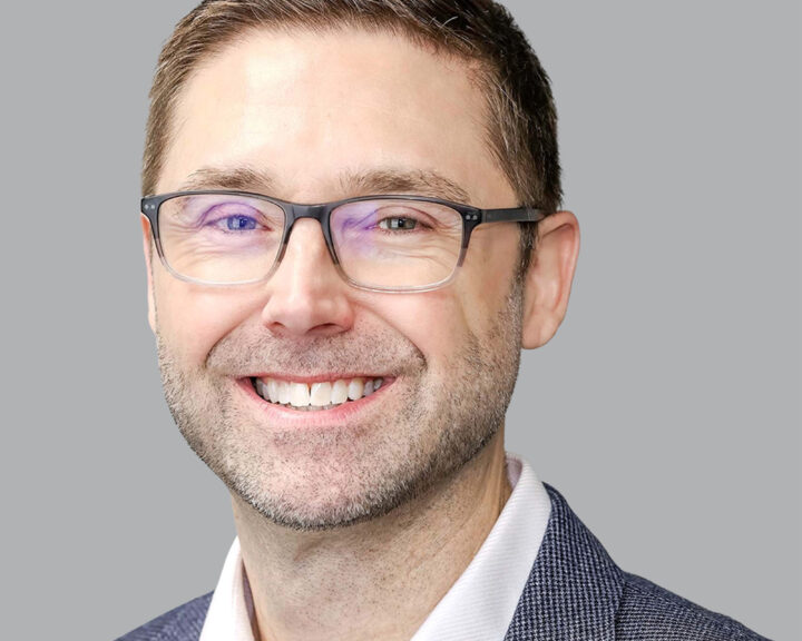 A man wearing glasses, a white shirt, and a textured blazer smiles at the camera against a plain grey background.