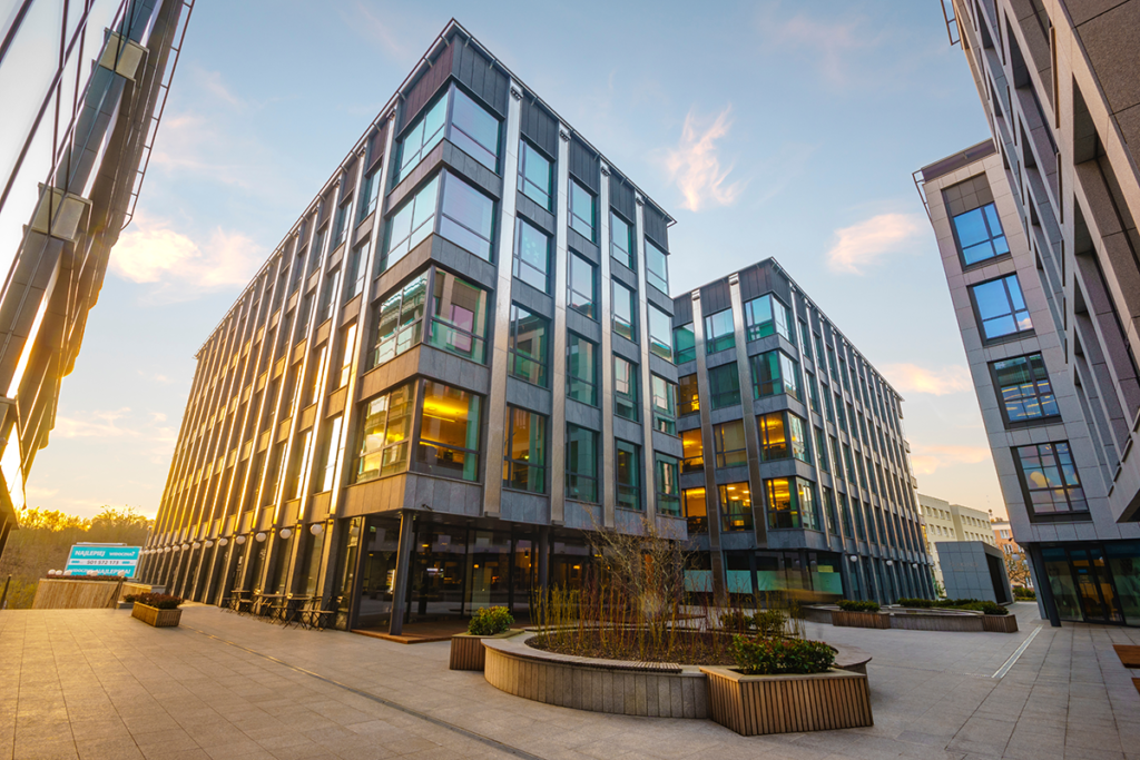 Modern office buildings with large glass windows and multiple stories, surrounded by paved walkways, planter boxes, and lit by the setting sun.