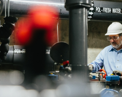 A worker in a hard hat operates machinery near labeled pipes in an industrial facility.