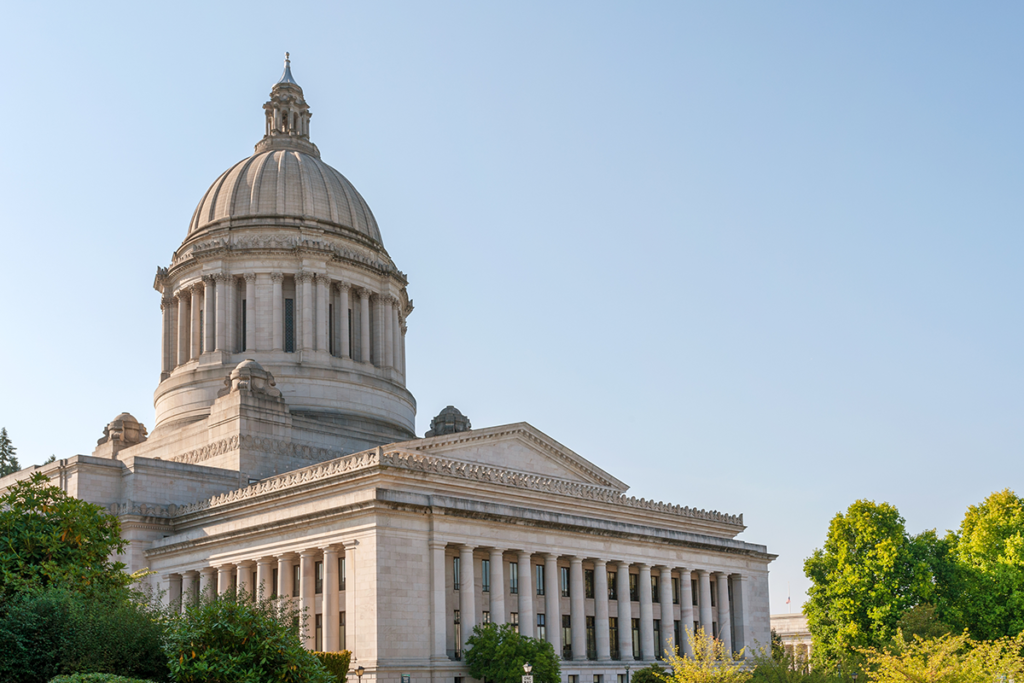 A large neoclassical government building with a domed roof and tall columns, surrounded by trees, under a clear blue sky.
